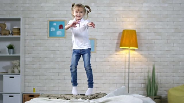 Slow motion shot of little schoolgirl with wavy golden hair and two ponytails jumping on bed, looking at camera and smiling