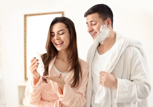 Young man with shaving foam and his girlfriend holding bottle of cream in bathroom