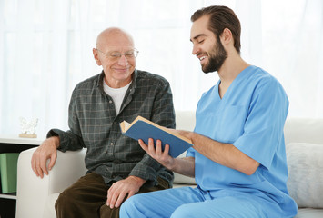 Young caregiver reading book to senior man at home