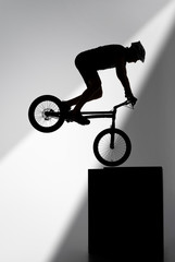 silhouette of trial cyclist performing stunt while balancing on cube on grey