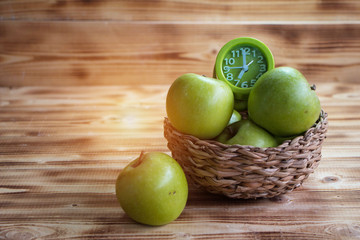The green apple and green alarm clock  in woven basket,put at the right side of wooden timber board,vintage warm light tone,blurry light around.