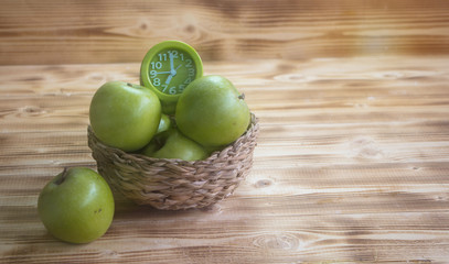 The green apple and green alarm clock in woven basket ,put on wooden timber boardvintage warm light tone,blurry light around.