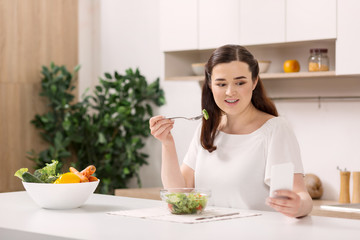 Bowl with vegetables. Pleasant attractive woman using smartphone and tasting salad