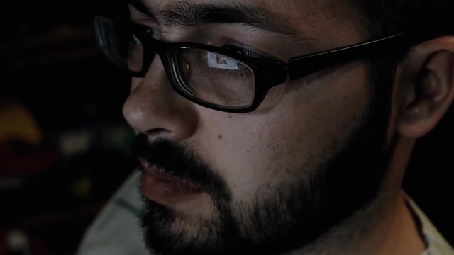 Portrait of a Bearded Young Man Wearing Glasses Sitting in His Office Working on a Computer. Computer Screen Reflects in His Glasses. Work on the computer at night