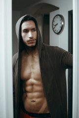sexy and naked muscular young man in jacket with hood standing near the window
