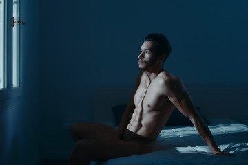 sexy and naked muscular young man sits on a bed in the room. the light from the window