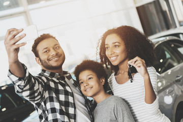Obraz premium African american family at car dealership. Mother, father and son are taking selfie in front of new car.
