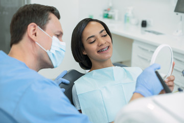 Amazing teeth. Smiling dark-haired patient looking in the mirror and her dentist sitting new her