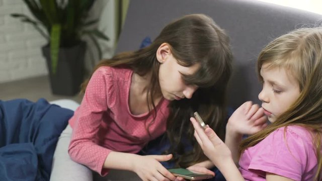 two sisters playing in gadgets in bed at home. dressed in pink pyjamas with a cellphone or smartphone. female kids is relaxing