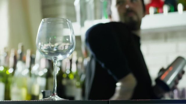 empty wineglass close-up stands on table and guy barman does tricks with bottles in unfocused background