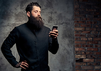 Portrait of a handsome bearded hipster dressed in a black shirt, using a smartphone, standing in a studio, isolated on a dark background.