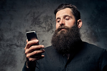 Portrait of a handsome bearded hipster dressed in a black shirt, using a smartphone, standing in a...