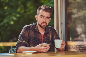A handsome fashionable male with stylish haircut and beard, wearing fleece shirt, drinking coffee and holding a smartphone in the cafeteria.