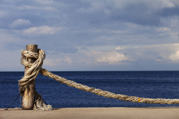 Obraz premium Rope knotted on wooden post in seafront, blue sea and cloudy sky