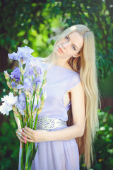 Attractive young girl with blonde dren hair and natural make-up smelling blue purple iris flowers on a background outdoors, tenderness and softness on a nature background