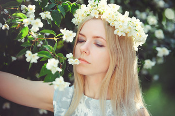 Attractive modest young girl with blonde with jasmine flowers wreath on head long hair and natural make-up in white dress outdoors, tenderness and softness on nature background