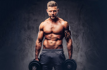 A handsome shirtless tattooed bodybuilder with stylish haircut and beard, wearing sports shorts, posing in a studio. Isolated on a dark background