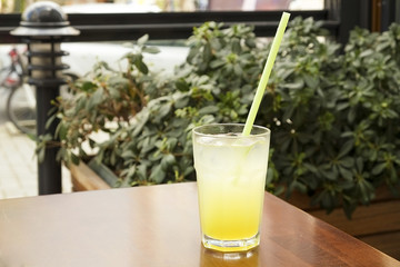 A glass of fresh ice cold lemonade on wooden table of coffee shop patio with green bushes on background. Close up of refreshing lemon juice cold beverage on the cafe terrace, outside. Text copy space