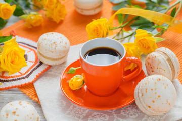 Orange cup of black coffee, yellow roses flowers and sweet pastel french macaroons
