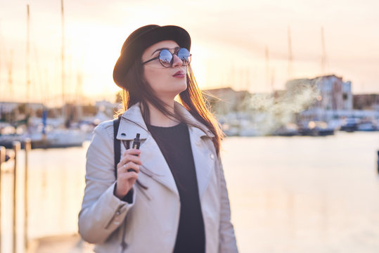 Pretty young woman in black hat with vape at a sea port