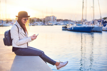 Fototapeta na wymiar Pretty young woman in black hat sitting on a pier with a phone at a sea port