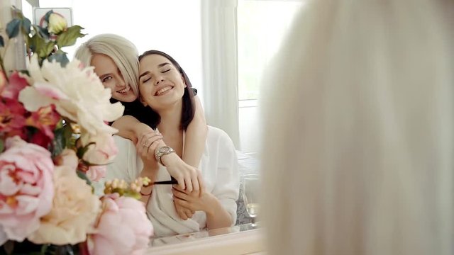 Brunette woman is doing makeup , looking in a mirror in bedroom. Her female friend is approaching back and embracing, both are smiling