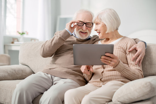 Pleasant rest. Delighted aged man fixing his glasses while looking at the tablet screen