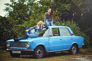 cheerful Attractive two young blonde girl and brunette posing on the hood of an old rusty car, dressed in jeans and shirts on a nature background