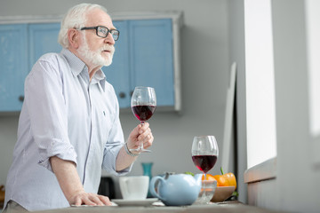 Lonely evening. Sad elderly man holding a glass of wine while looking into the window