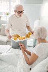 Tasty breakfast. Pleasant nice senior man holding a tray with food while talking to his wife