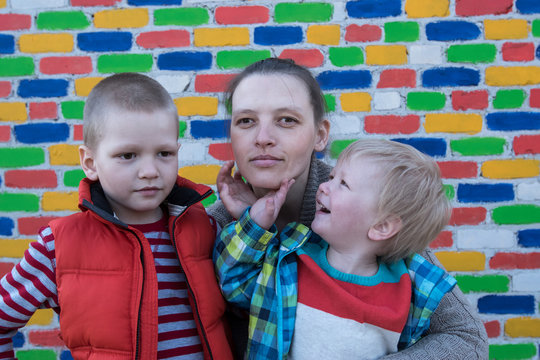 Happy mother hugs her two charming sons, blond and shaten, against the backdrop of multicolored brick wall. Caucasian children and mom have good time together.