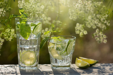 two glasses with a mojito drink in the open air, next to lie lime slices, against a background of greenery, sun rays
