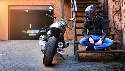 Lonely biker thought about and rested on the stairs near the motorcycle. Motorbike and motorcyclist in a helmet on a city street.