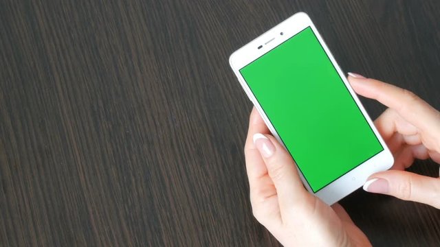 Female hands with beautiful French manicure take a white smartphone with Green Screen. Using Smartphone,Holding Smartphone with Green Screen on a stylish black wooden table