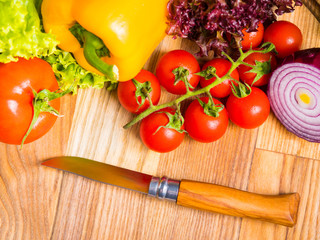Appetizing still life. Fresh bulgarian yellow pepper, cherry tomatoes, lettuce leaves, red onion, green basil on wooden board. The knife for cutting vegetables.