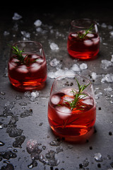 Pomegranate cocktail with ice and rosemary surrounded with crushed ice on the black concrete background.