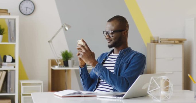 Young African American man in glasses and jeans shirt sitting at the laptop computer at work when taping and chatting on the smartphone and making dissapointed face. Inside