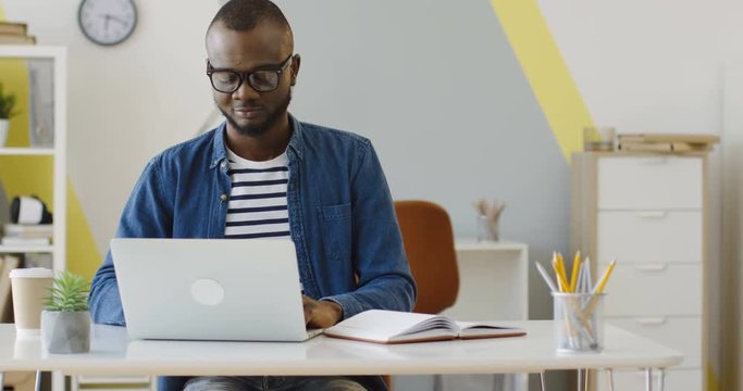 African American young male office worker in the glasses and jeans shirt working, typing and thinking in the nice urban office. Inside