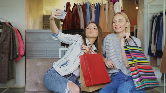 Attractive female friends are using smartphone to make selfie while sitting in clothing shop with colourful paper bags. They are smiling, posing, laughing, gesturing.