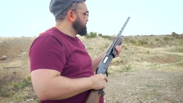 Overweight hunter fires a shotgun and ejects a smoking shell from the chamber in slow motion. Bearded man aims his firearm at a flying target, pulls the trigger, and removes the cartridge.