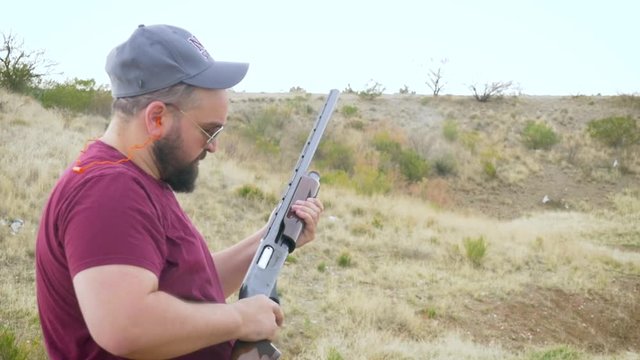 Young man shoots an orange clay pigeon with a pump action shotgun in slow motion. Overweight man in a baseball hat at a desert gun range aims his firearm at the flying target and pulls the trigger.