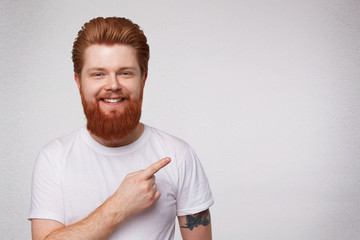 Indoor shot of young excited surprised redhead guy with beard point with a finger aside, smiles and feels happy