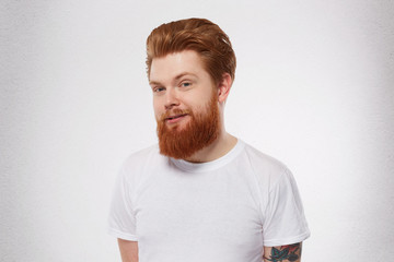 Obraz premium Young redhead man with heavy, massive beard smiles and feels happy. Portrait of pleased, joyful male. Isolated over studio white wall