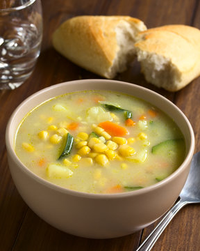 Vegetarian corn and courgette chowder served in bowl, glass of water and bread roll in the back, photographed on dark wood with natural light (Selective Focus, Focus in the middle of the soup)