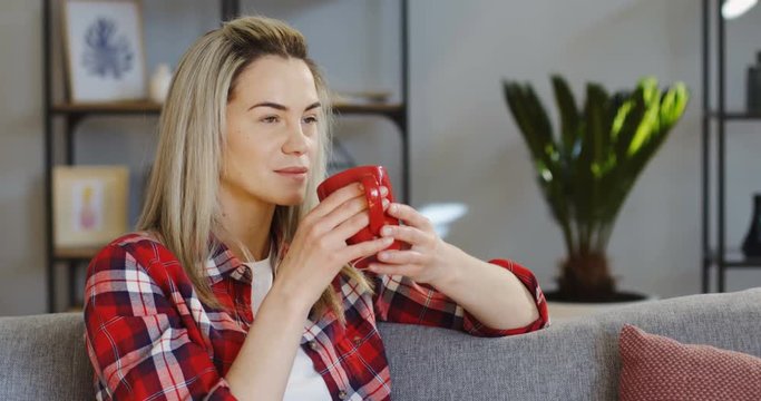 Beautiful blonde woman in the motley shirt drinking hot tea or coffee from a red cup on the gray sofa in the living room. Indoor
