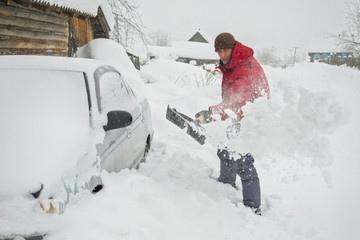 a man in a red jacket cleans the snow near his gray car to release his snow blockage