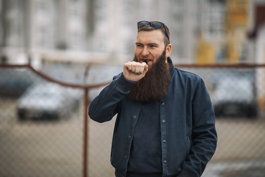 Vape bearded man in real life. Portrait of young guy with large beard in glasses vaping an electronic cigarette opposite urban background in the spring.