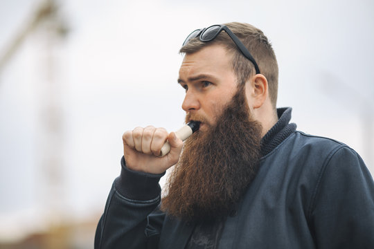Vape bearded man in real life. Portrait of young guy with large beard in glasses vaping an electronic cigarette opposite urban background in the spring.