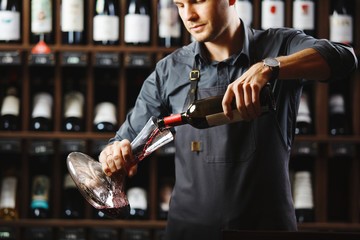 Bartender pours red wine in transparent vessel in cellar