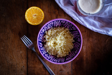 Bowl of couscous in purple turkish ceramic bowl on a wooden tabletop with natural light, orange in half an ice tea